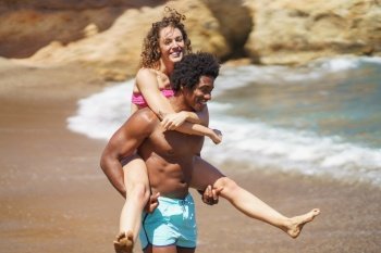 Happy young multiracial couple fooling around on sandy beach near waving ocean while enjoying summer vacation together during sunny weekend. Joyful black man giving piggyback ride to girlfriend near sea