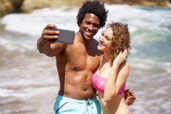 Happy young multiethnic couple in swimwear hugging while taking selfie on mobile phone for social media during honeymoon. Smiling diverse couple taking selfie near sea