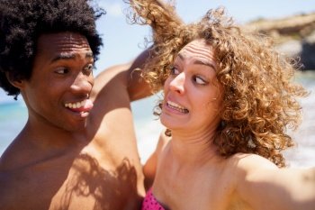 Cheerful black man with Afro hair and tongue out touching curly hair of girlfriend making funny grimace on sunny day near sea. Selfie of funny diverse couple making faces on beach