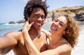 Cheerful young woman pulling smile on black boyfriend with Afro hair by stretching corners of mouth while taking selfie on beach during summer vacation. Selfie of happy woman pulling smile on boyfriends face
