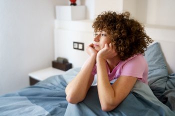 Hispanic curly woman leaning on arms and looking away thoughtfully while sitting on bed after getting up. Dreamy lady sitting on bed in morning