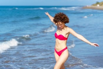 Delighted young fit female with dark curly hair, in red bikini smiling and looking down while walking in wavy sea with outstretched arms during summer holidays holidays. Joyful young lady in swimwear walking in sea with outstretched arms