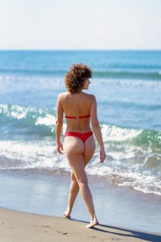 Back view of gorgeous young fit female with brown curly hair in red bikini, smiling and admiring wavy sea while strolling on sandy beach on sunny day. Calm woman walking on sandy seashore during summer vacation