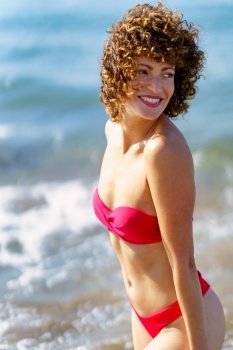 Side view of excited young female tourist with curly hair in stylish red bikini, smiling and looking over shoulder while standing on beach during summer vacation. Graceful happy lady standing on seashore and smiling
