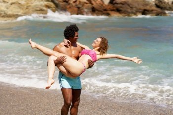 Happy African American man raising woman with outstretched hands in swimwear while looking at each other during vacation against waving sea. Cheerful multiethnic couple having rest together on seashore