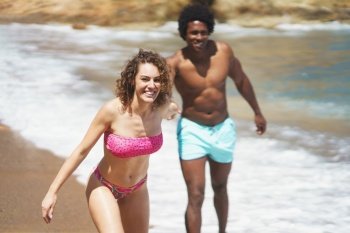 Cheerful multiethnic couple in colorful swimwear running on sandy shore while holding hands having fun together on resort. Joyful multiracial couple fooling around on beach