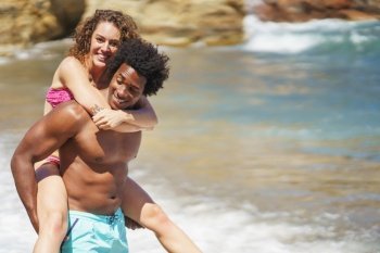 Cheerful young African American man with Afro hair giving piggyback ride to happy girlfriend on beach during honeymoon. Black man carrying girlfriend near sea