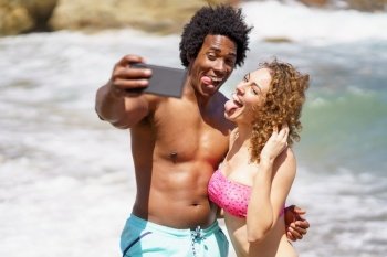 Cheerful multiracial couple in swimwear hugging and showing tongues while taking selfie on mobile phone for memory against waving ocean. Funny multiethnic couple showing tongues while taking selfie on beach