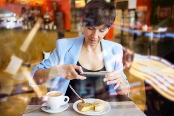 Through glass view of young female in blue jacket, sitting at table with coffee cup and looking at screen of mobile phone while taking picture of snack in restaurant. Young woman taking photo of dish with smartphone