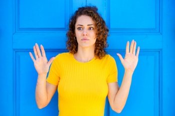 Afraid young female in yellow t shirt with curly brown hair and raised hands looking away against blue wall on street. Scared woman with hands up on blue background
