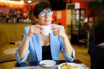 Thoughtful middle aged female in blue jacket and eyeglasses sitting at table with cup of hot beverage and looking away while resting in cafe. Pensive mature woman drinking coffee in cafe