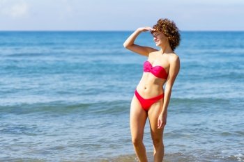 Side view of smiling young female with curly red hair, in bikini standing in seawater with hand at face covering sunlight while looking away against blue wavy sea. Slim woman in bikini standing in seawater