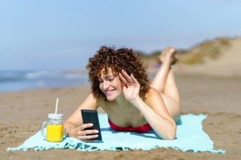 Young happy female with ginger curly hair lying on sandy shore with orange juice and waving hand while having video call on phone. Smiling woman chatting on mobile phone on beach
