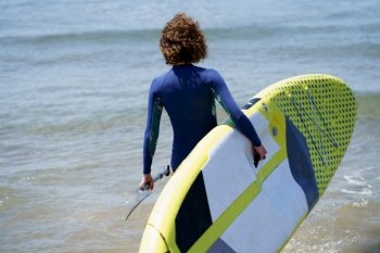 Back view of unrecognizable female surfer in wetsuit with SUP board and paddle standing in water at beach in rippling blue sea at daylight. Unrecognizable surfer with paddleboard in sea