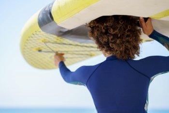 Back view of unrecognizable curly haired female in blue swimwear lifting white yellow SUP board on head while facing blue sea in bright daylight. Unrecognizable woman lifting paddleboard over head on shore