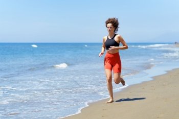 Concentrated barefoot young female in sportswear and with curly hair looking away, while jogging in daylight on sandy beach near foamy seawater against blurred blue sky. Focused young woman exercising on beach near water