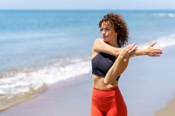 Pensive young sportswoman with curly hair in sportswear stretching arm while standing near waving sea and looking away. Dreamy woman stretching near sea