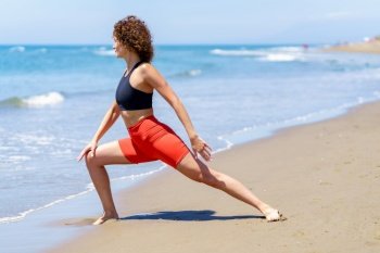 Full body side view of curly haired female athlete in activewear doing lunges near waving sea on sunny day. Fit woman training on sandy beach