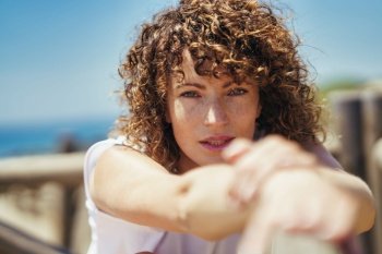 Young curly haired female with freckles and brown wavy hair in white casual clothes stretching arms while leaning on fence against blurred background. Young woman stretching arms on bridge