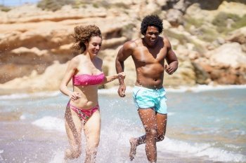 Young multiracial couple in swimwear running in splashing water while looking down and laughing together during sunny day near sea waves. Happy diverse couple sprinting in seawater and enjoying