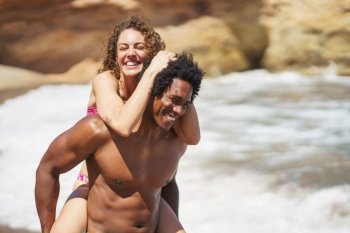 Joyful young African American man giving piggyback ride to cheerful curly haired woman having fun on seashore during sunny vacation laughing and smiling. Cheerful woman holding on to boyfriends head while having piggyback ride