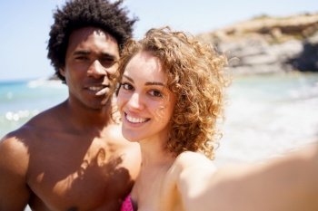 Delighted young woman outstretching hand towards camera while taking selfie with black boyfriend against sea and rocky cliff. Selfie of happy diverse couple on beach
