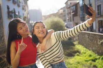 Happy Asian woman showing horn gesture and sitting with hand on shoulder of friend while taking self portrait on smartphone together in Granada. Young Asian female friends taking self portrait on cellphone during sightseeing