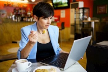 Concentrated young female in blue jacket with short hair holding snack in spoon and sitting at table with coffee while looking at screen of laptop in restaurant. Focused young woman using laptop in cafe