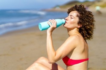 Side view of young redhead female in bikini bra drinking refreshing water from bottle while sunbathing on coast. Woman drinking water on sunny beach