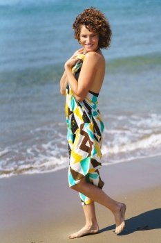 Full body side view of positive young female in colorful dress standing on sandy beach near waving sea in daylight and looking at camera. Smiling woman standing on sandy beach