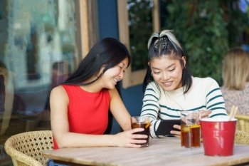 Positive young Asian female friends, in casual outfits sitting at wooden table with glasses of cold drinks and looking at smartphone screen together while watching video during meeting. Cheerful Asian women sharing smartphone at table in cafe