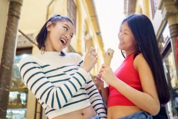Positive Asian woman friends eating tasty ice cream while standing on street and enjoying time together in daytime. Smiling young Asian women with ice creams