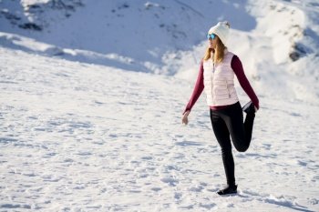 Full body of young female traveler in cozy clothes and beanie cap standing on one leg and holding leg up while looking away on snowy slope in daylight. Young sporty woman standing on one leg on snowy terrain