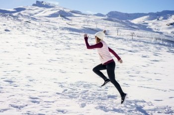 Side view of unrecognizable female traveler in warm clothes and beanie running on snowy slope of mountainous terrain in winter with blue sky. Excited young woman running on snowy plain
