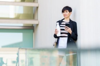 Serious businesswoman in formal clothes with handbag and smartphone leaning on hand bottle of drink while standing near glass railing. Pensive young woman with smartphone and hand bottle