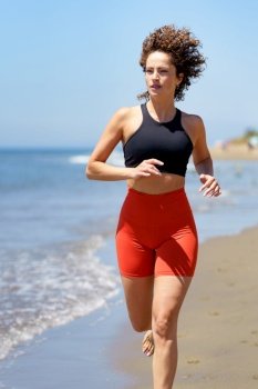 Smiling curly haired barefoot young female in sportswear warming up and looking away while jogging alone on sandy beach near waving sea against blurred blue sky. Happy young woman enjoying running exercise on beach