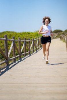 Full body of young female athlete in sportswear running along wooden path during fitness training on sunny summer day. Confident sportswoman jogging on wooden path