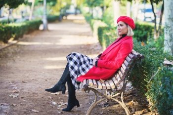 Full body side view of female in red outfit and beret sitting on wooden bench in autumn park with green plants. Stylish woman on bench in autumn park