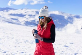Smiling young female traveler in warm clothes beanie wool cap polarized sunglasses, looking down and opening thermos water bottle while standing on snowy mountain slope in sunny winter day. Happy woman standing with water bottle in sunlight on snowy terrain