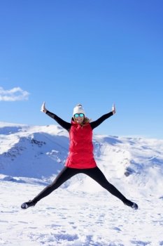 Full body of excited young female in vest beanie cap polarized sunglasses looking at camera, while leaping in air with widened legs stretched arms against snowy mountains and blue sky. Carefree young woman jumping with raised arms in snowy terrain