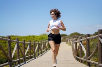 Full body of young fit female athlete in sportswear jogging on wooden boardwalk while warming up during fitness workout. Sportswoman running along wooden path on sunny day