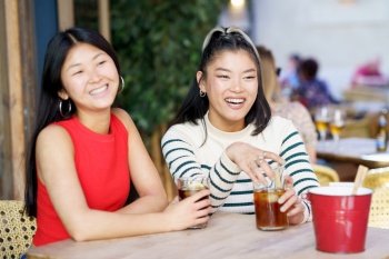 Happy young Asian women in casual outfits smiling and looking away while sitting at wooden table with refreshing cold beverages during meeting together. Positive Asian girls resting in cafe with drinks