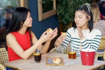 Delighted young Asian female friends in casual wear eating delicious food with closed eyes while sitting at table with cold beverages in cafe together. Satisfied Asian women eating tasty meal in cafe together
