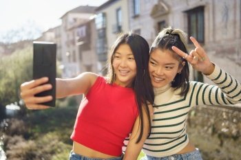 Happy Asian girlfriends in casual clothes smiling and looking at screen while making selfie on mobile phone and showing rock gesture during daytime in Granada. Positive Asian female tourists shooting selfie on cellphone