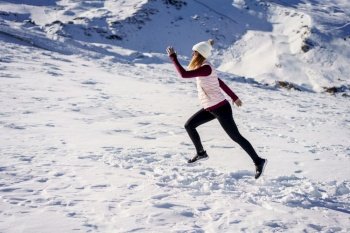 Full body side view of active female in sportswear and beanie jumping on white snow in winter while training at daylight. Active woman jogging on snowy terrain