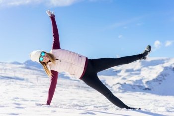 Full body of positive young female in cozy clothes and stylish sunglasses, stretching arms in side plank yoga pose with open legs on snowy ground against blue sky. Happy woman with eyeglasses in side plank yoga handstand pose