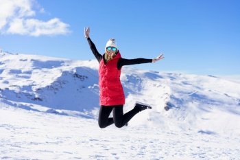 Happy young female in warm clothes cap polarized sunglasses looking at camera, while jumping in air with bent legs and stretched arms in daylight against snowy mountains and blue sky. Carefree young woman jumping with raised arms on snow terrain