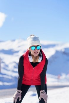Young healthy woman in outerwear and sunglasses resting while leaning on knees against mountains in snow. Female runner in warm clothes in winter