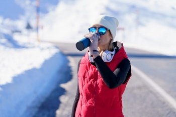 Sportswoman in warm clothes and sunglasses drinking from thermos bottle while having workout in snowy mountains. Athletic woman in outerwear drinking water in snowy terrain