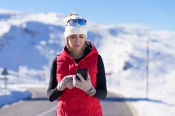 Adult athletic female in warm outerwear browsing smartphone while standing on asphalt road in winter mountains. Sportive woman using phone in winter countryside